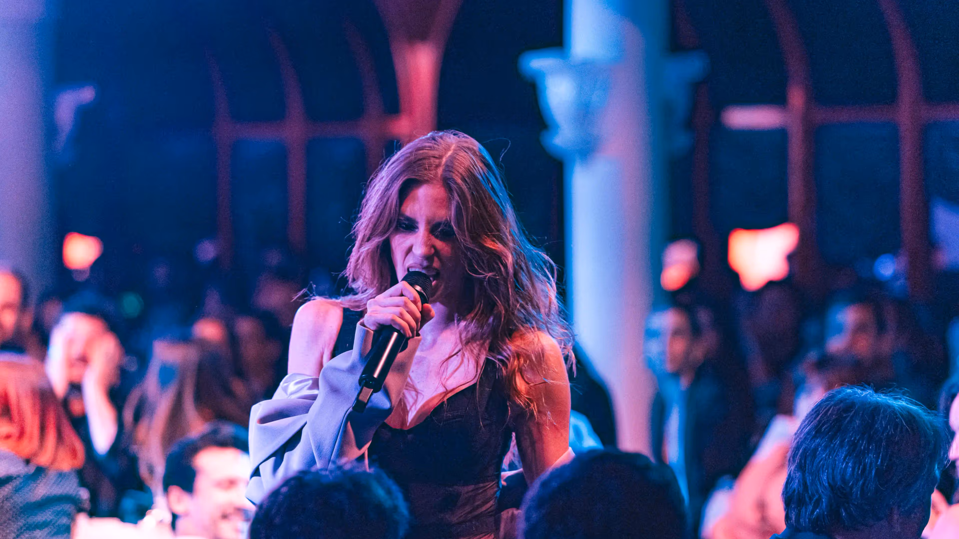 A woman sings into a microphone in front of an audience in a room with colorful lights.