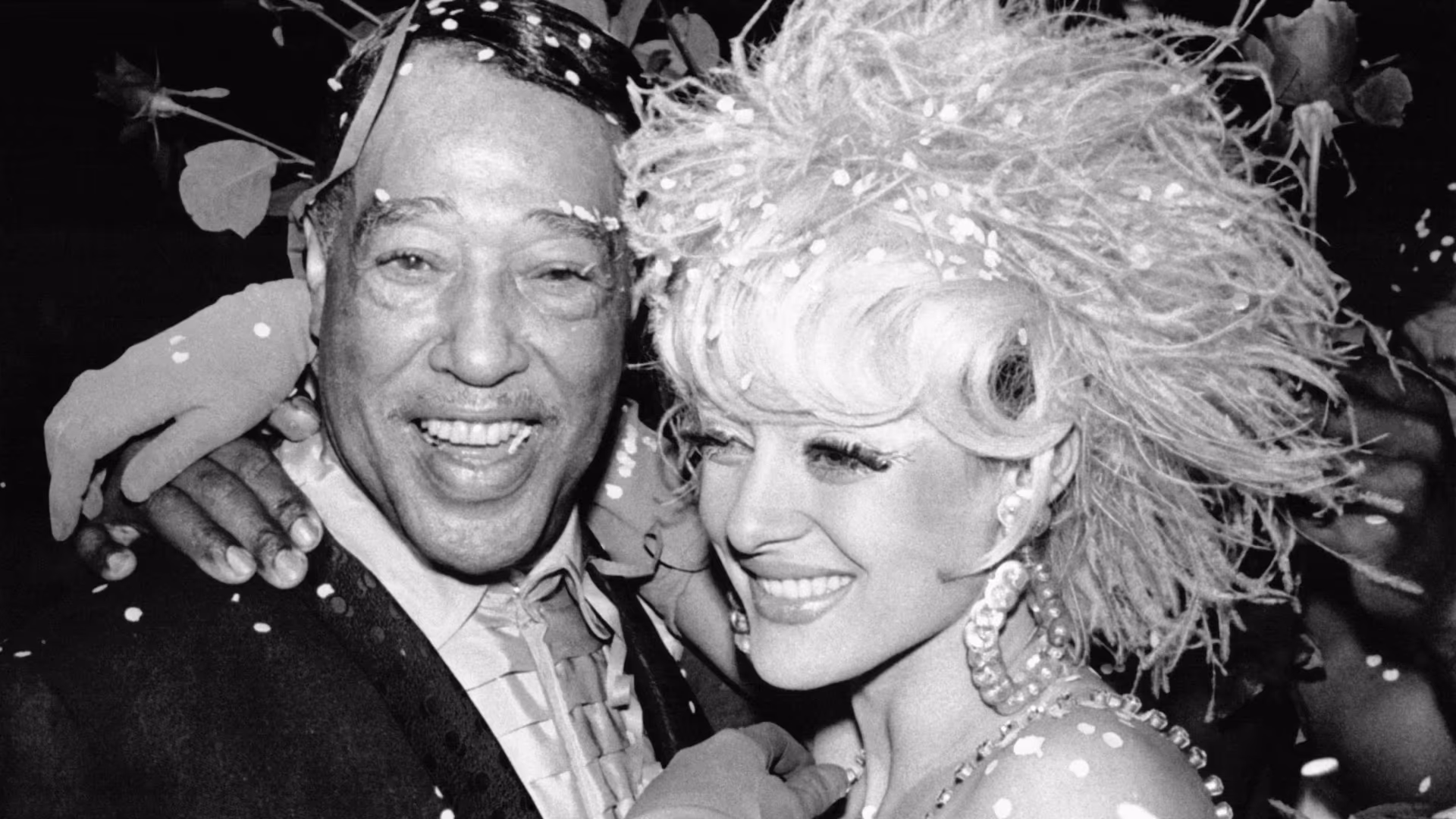 Two smiling people wearing festive outfits with accessories and confetti around them