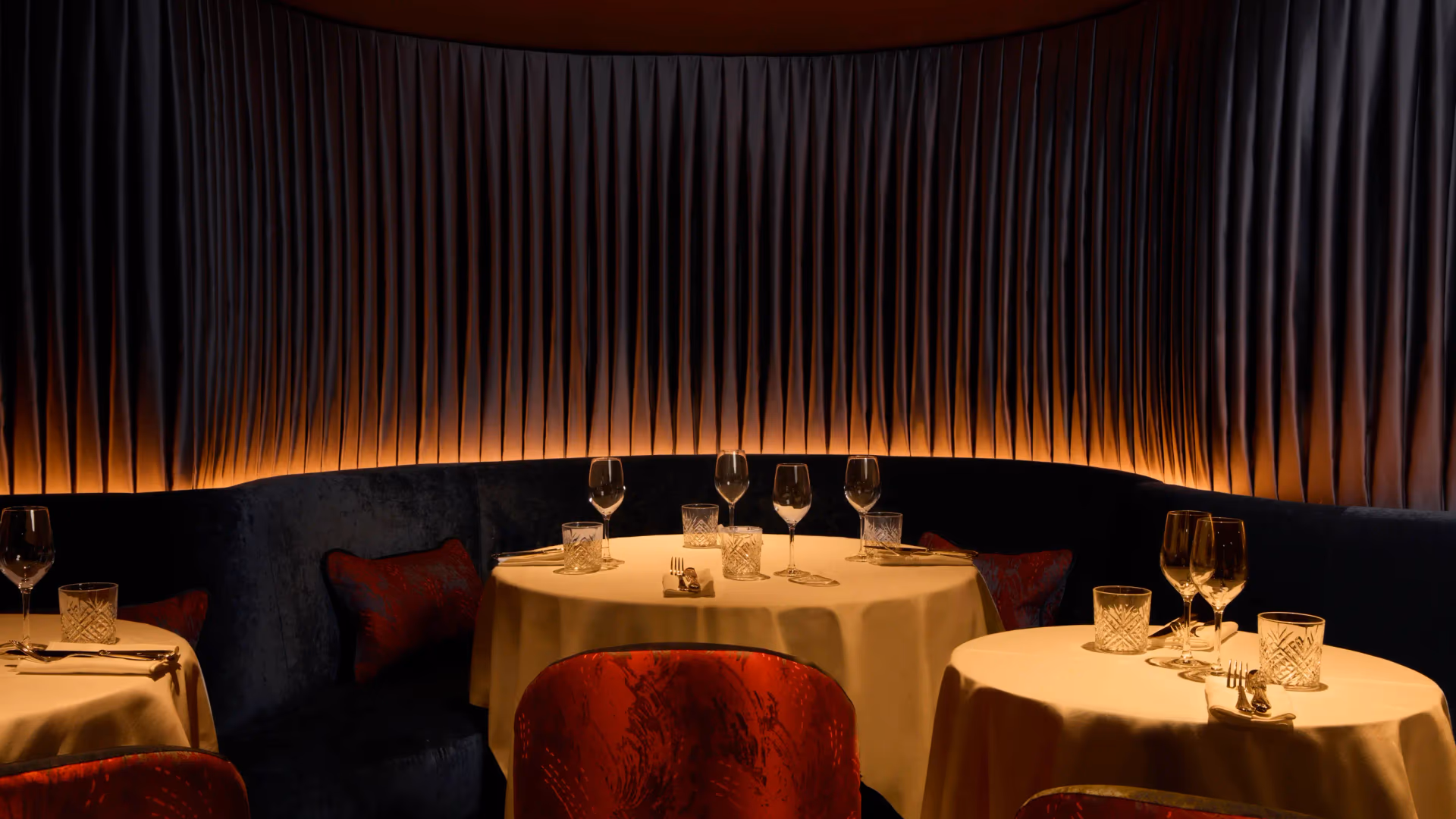 A round table with glasses and cutlery, surrounded by red chairs, in a dimly lit restaurant with dark curtains.