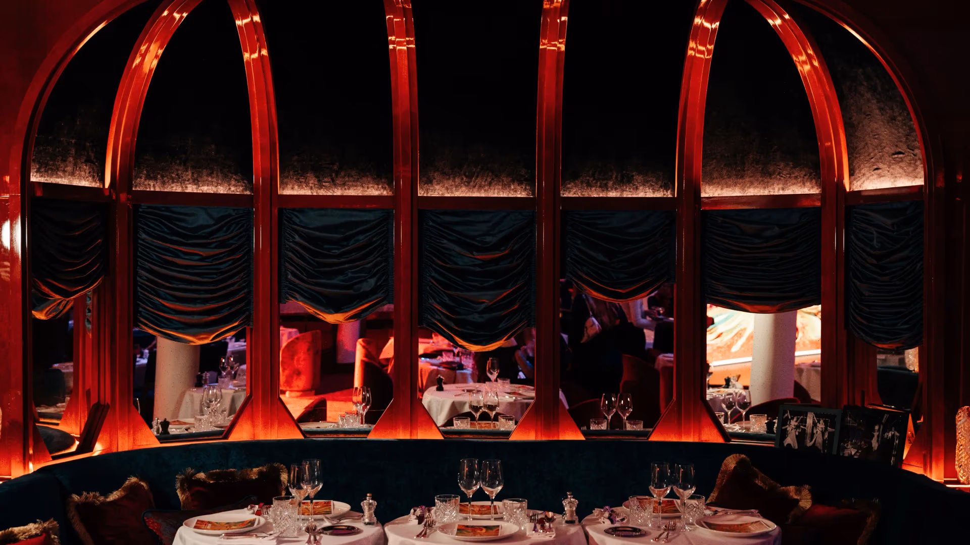 Three round tables with glasses and cutlery are arranged in front of a large red-arched window in a dark and elegant interior.
