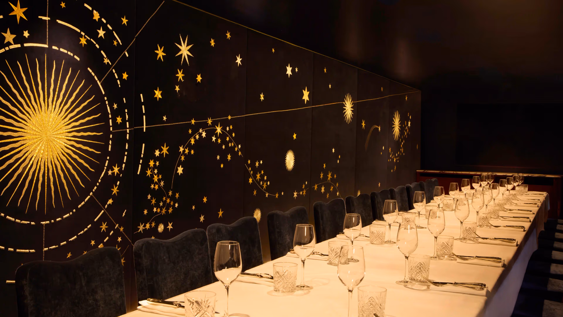 A long table set with glasses and cutlery, surrounded by chairs, in a room decorated with glowing star patterns on a black background.